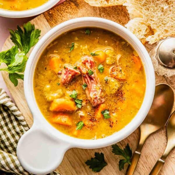 A bowl of hearty soup with chunks of meat and vegetables, garnished with herbs, served alongside slices of bread and a set of spoons on a wooden board.