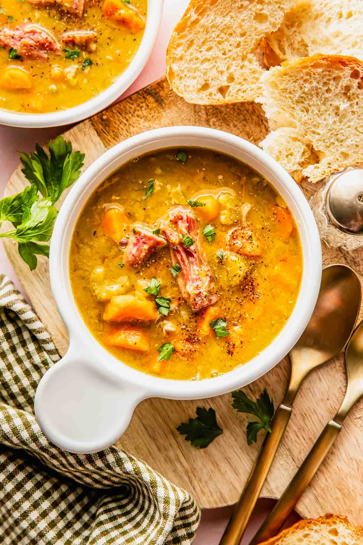 A bowl of hearty soup with chunks of meat and vegetables, garnished with herbs, served alongside slices of bread and a set of spoons on a wooden board.