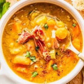 A bowl of split pea soup with shredded ham, carrots, and herbs, served with a spoon on a wooden surface next to bread slices and parsley.