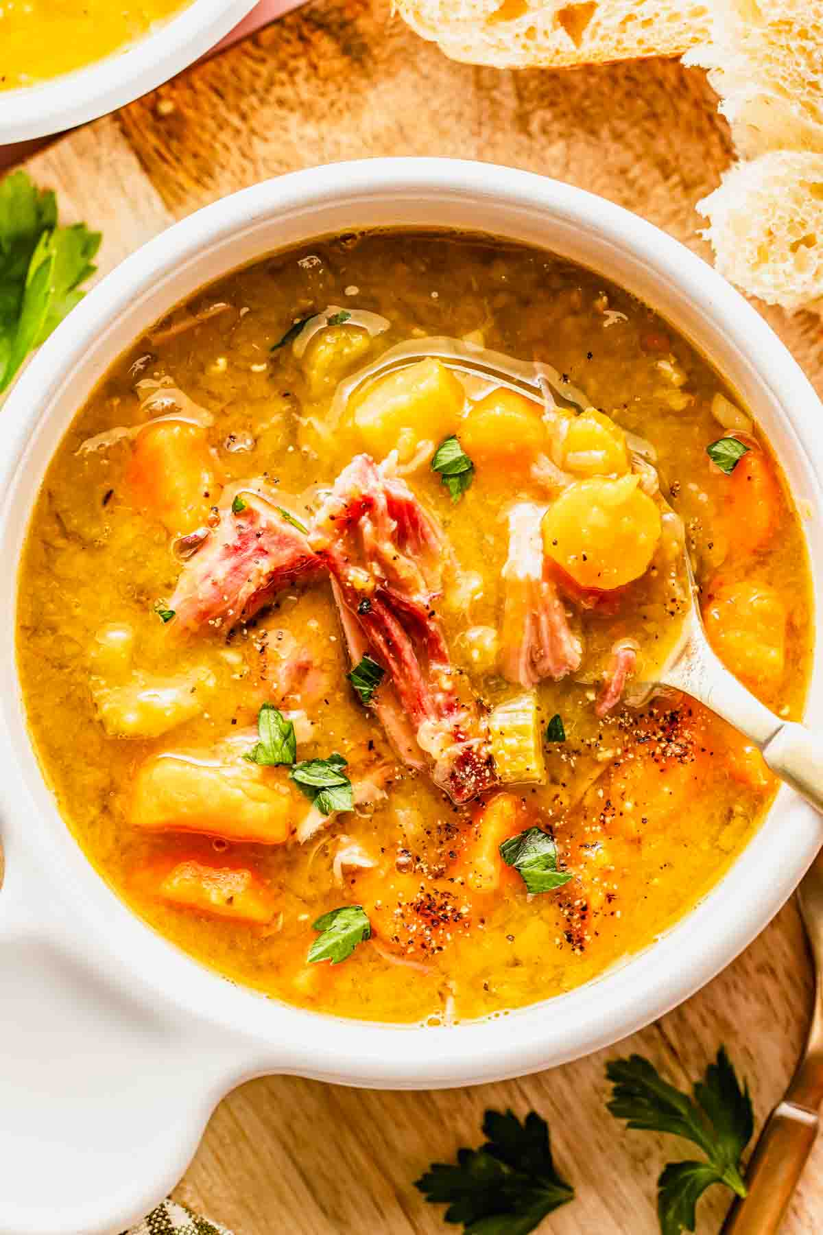 A bowl of split pea soup with shredded ham, carrots, and herbs, served with a spoon on a wooden surface next to bread slices and parsley.