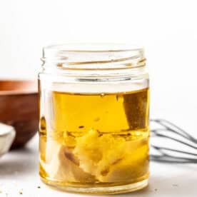 A glass jar containing oil and mustard, with a whisk and wooden bowl in the background on a white surface.