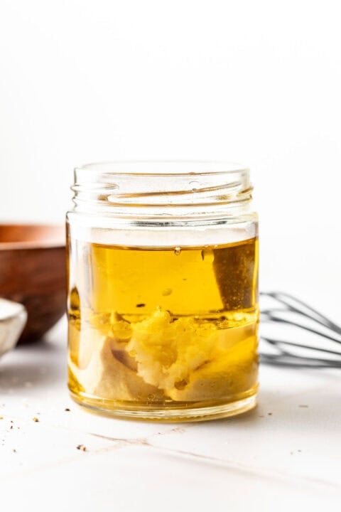 A glass jar containing oil and mustard, with a whisk and wooden bowl in the background on a white surface.