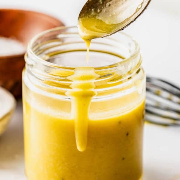 A spoon drips yellow salad dressing into a glass jar, with a whisk and a wooden bowl in the background on a white surface.