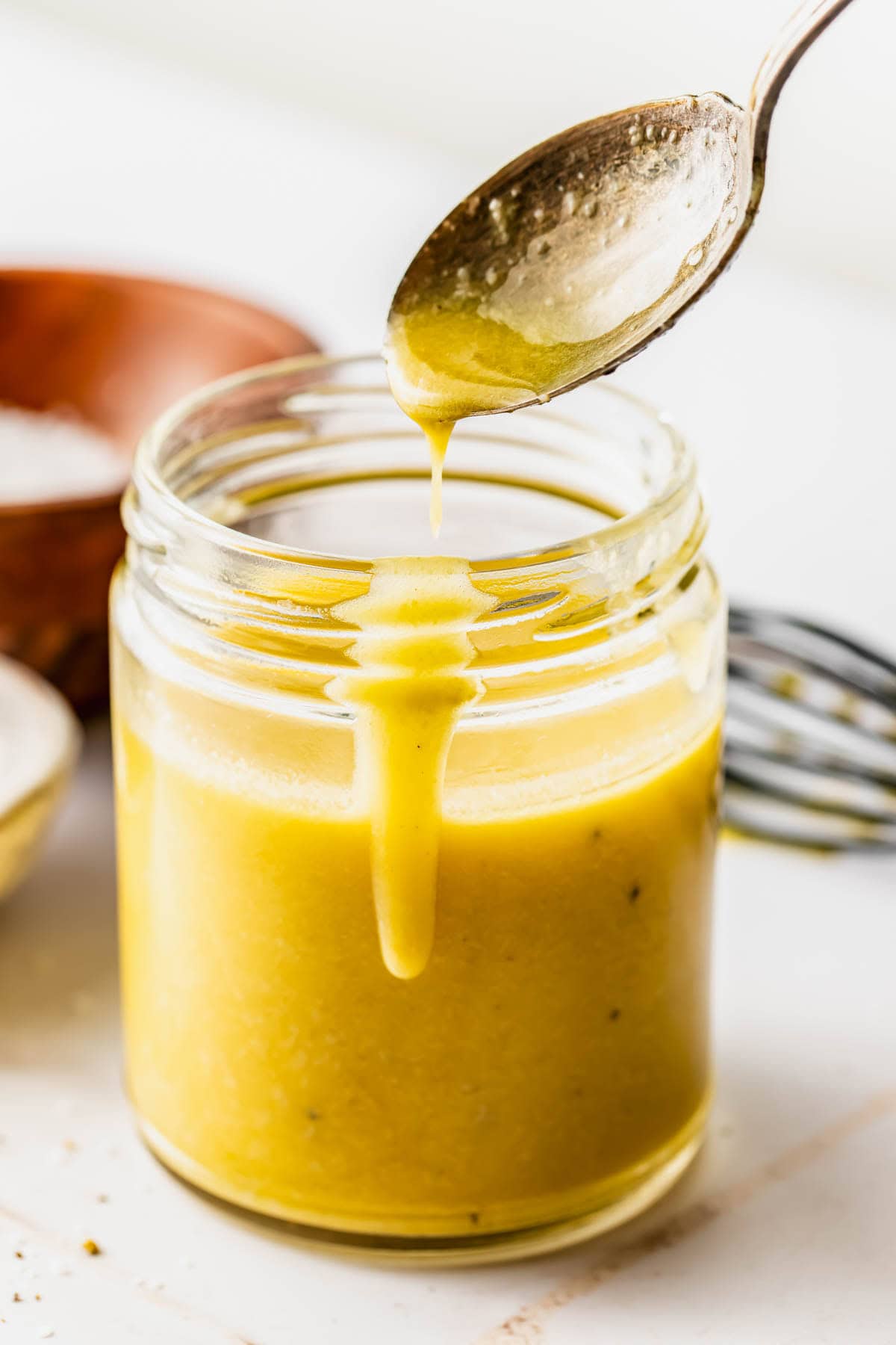 A spoon drips yellow salad dressing into a glass jar, with a whisk and a wooden bowl in the background on a white surface.