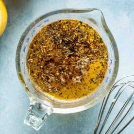 A glass measuring cup with a herb and oil mixture next to a metal whisk and sliced lemons on a blue surface.