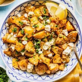 A bowl of roasted potatoes topped with crumbled feta cheese, chopped herbs, and two lemon wedges, with a spoon and garnishes nearby.