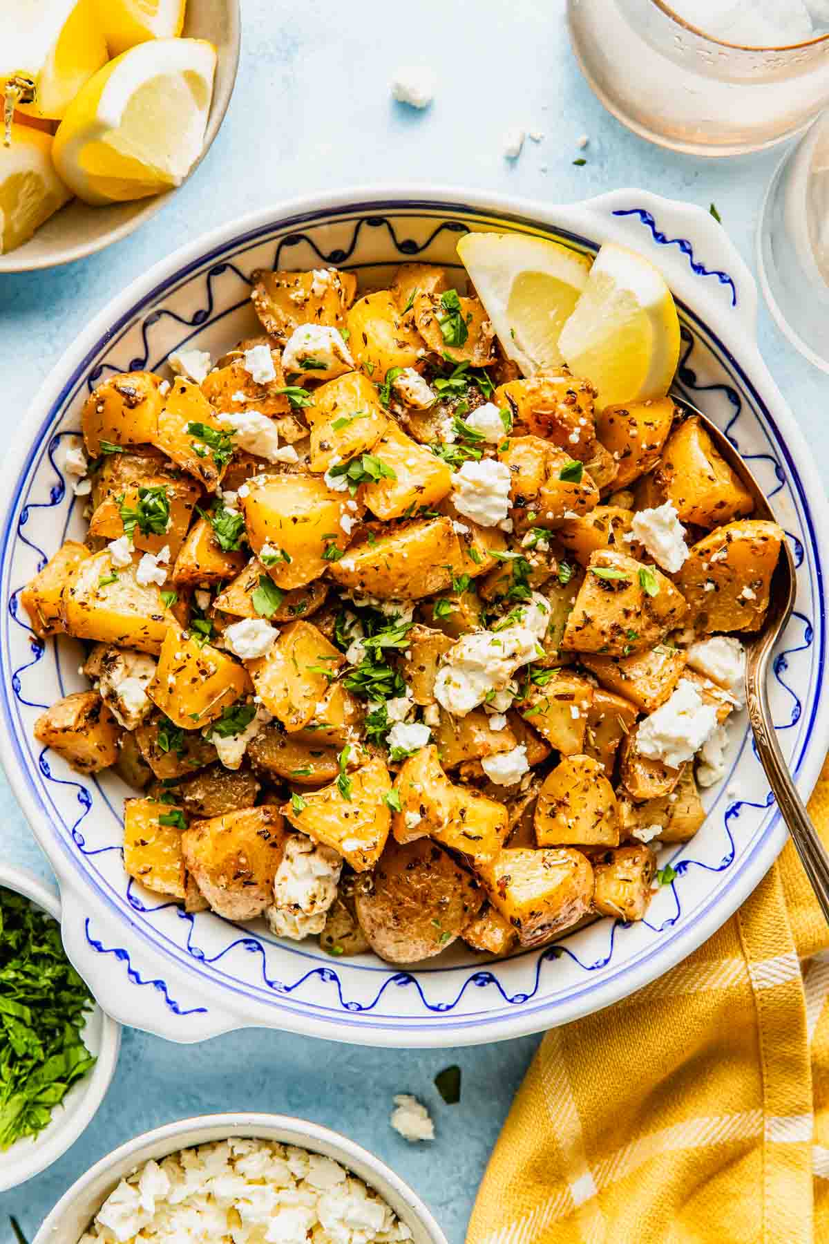 A bowl of roasted potatoes topped with crumbled feta cheese, chopped herbs, and two lemon wedges, with a spoon and garnishes nearby.
