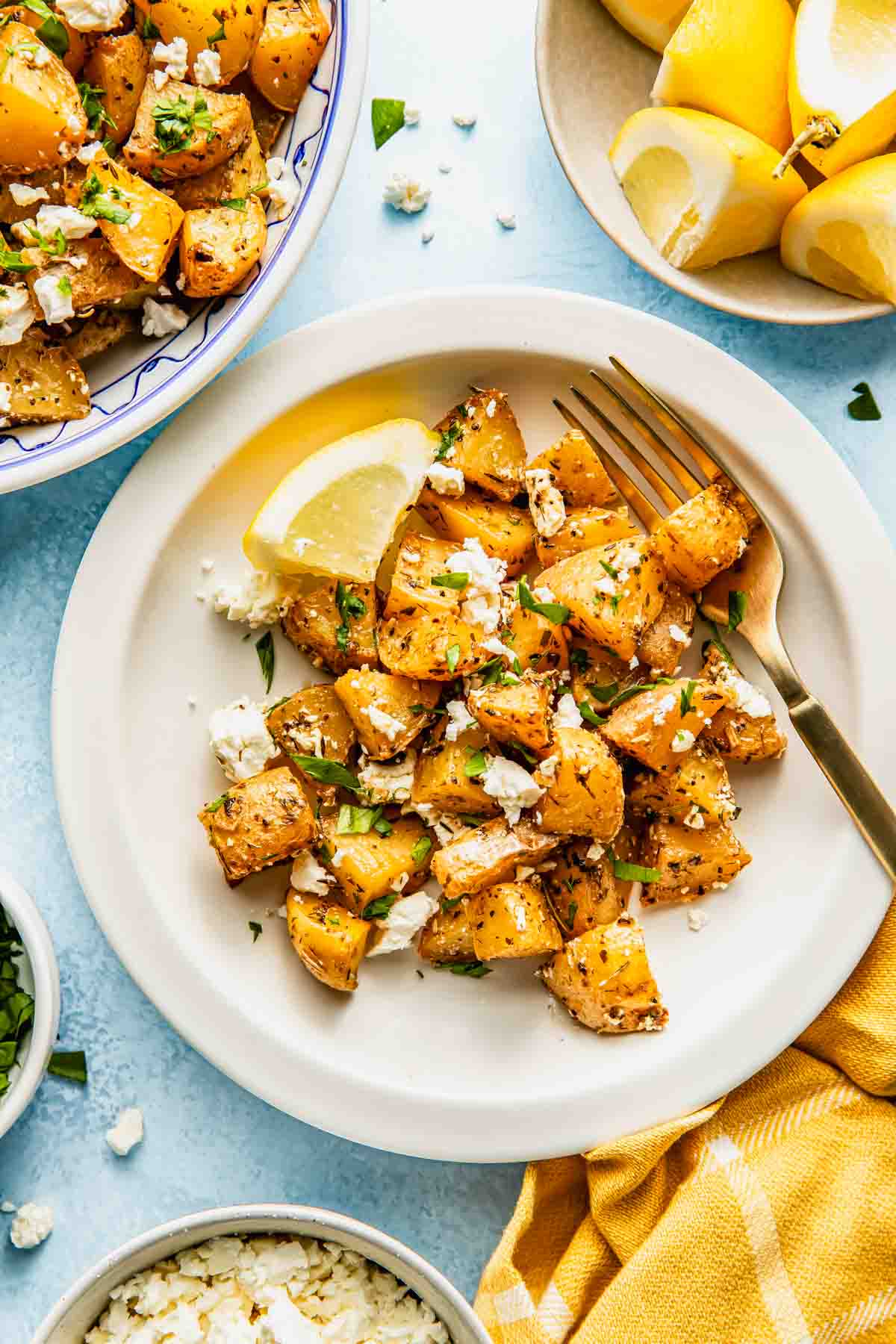 A plate of roasted potatoes garnished with crumbled feta cheese and herbs, served with a lemon wedge and a fork. Additional potatoes, lemon wedges, and cheese are nearby.