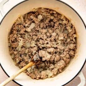 Ground beef and chopped onions being cooked in a white pot, stirred with a wooden spoon.