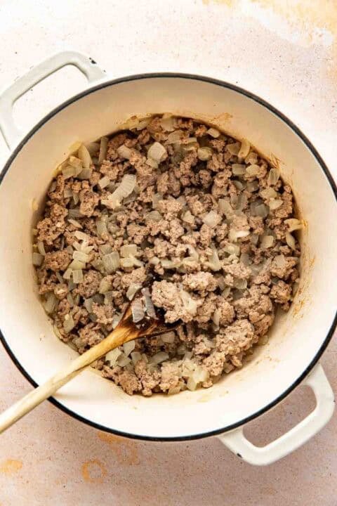 Ground beef and chopped onions being cooked in a white pot, stirred with a wooden spoon.