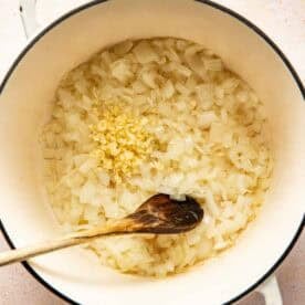 Diced onions and minced garlic being sautéed in a white pot with a wooden spoon.