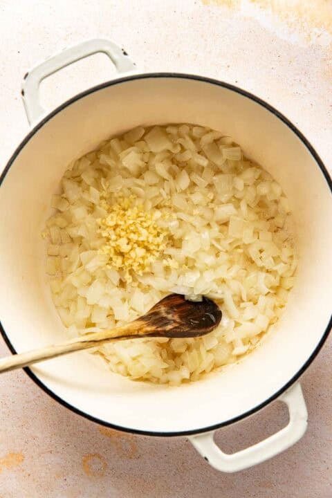 Diced onions and minced garlic being sautéed in a white pot with a wooden spoon.