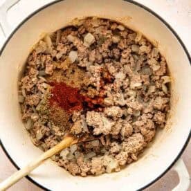Ground turkey and chopped onions cooking in a white pot with various spices on top, stirred with a wooden spoon.