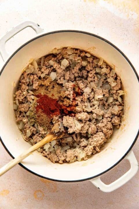 Ground turkey and chopped onions cooking in a white pot with various spices on top, stirred with a wooden spoon.