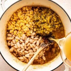 A pot containing white beans, corn, green chilies, and broth being poured in, with a wooden spoon resting inside.