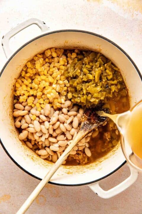 A pot containing white beans, corn, green chilies, and broth being poured in, with a wooden spoon resting inside.