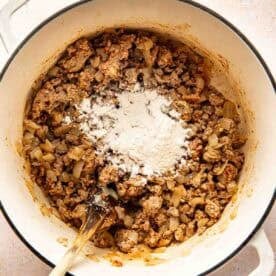A white pot containing cooked ground meat and onions with a mound of flour on top, being stirred with a wooden spoon.