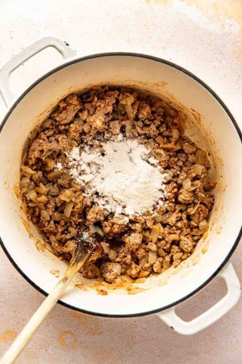A white pot containing cooked ground meat and onions with a mound of flour on top, being stirred with a wooden spoon.