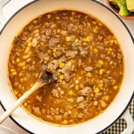 A pot of chili with ground meat, corn, beans, and broth is being stirred with a wooden spoon; bowls of chopped cilantro and diced avocado are nearby.