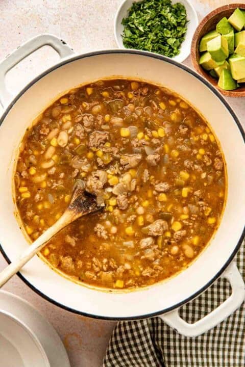 A pot of chili with ground meat, corn, beans, and broth is being stirred with a wooden spoon; bowls of chopped cilantro and diced avocado are nearby.