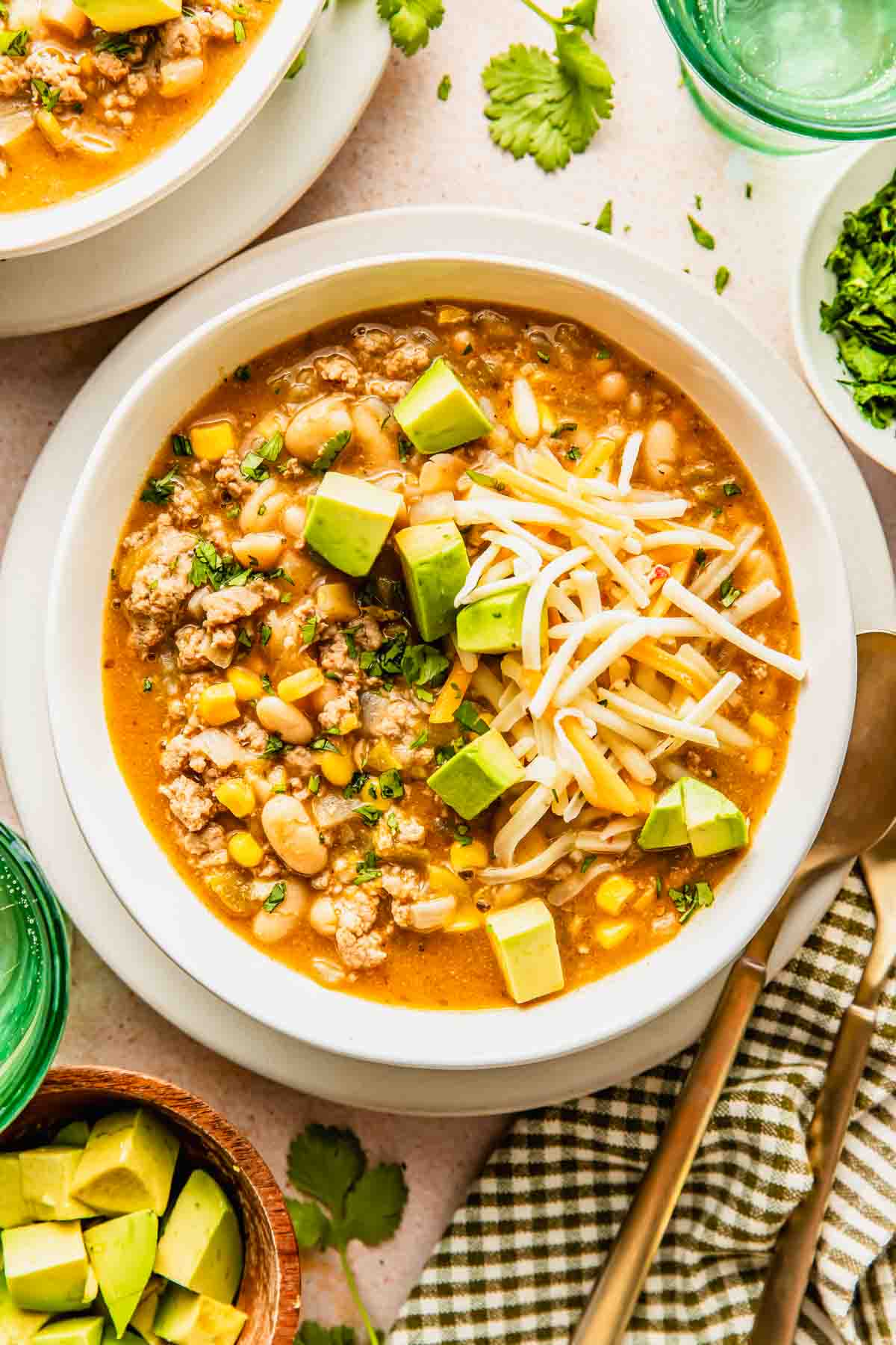 A bowl of white chili topped with shredded cheese and diced avocado, surrounded by bowls of fresh cilantro and avocado, with a fork and spoon on the side.