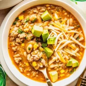 A bowl of white chili with ground turkey, corn, beans, shredded cheese, avocado cubes, and herbs, served with a spoon.