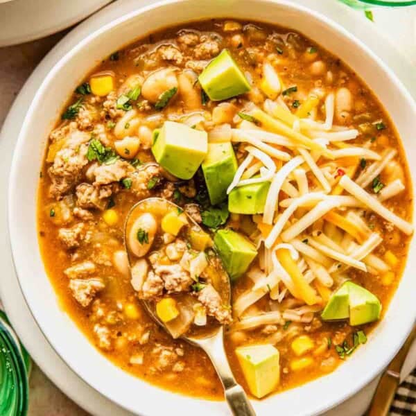 A bowl of white chili with ground turkey, corn, beans, shredded cheese, avocado cubes, and herbs, served with a spoon.