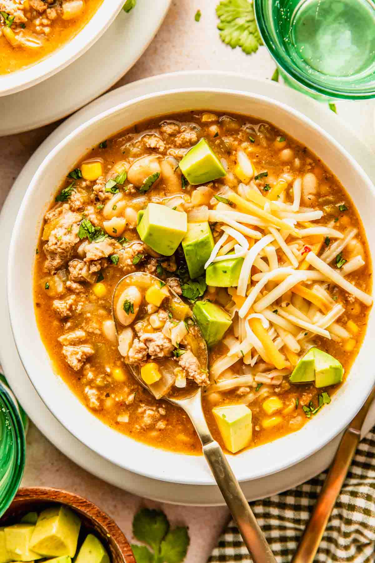 A bowl of white chili with ground turkey, corn, beans, shredded cheese, avocado cubes, and herbs, served with a spoon.