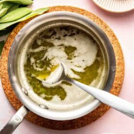 A saucepan with melted butter and foam, stirred by a spoon, sits on a cork mat. Salt, pepper, sage leaves, and breadcrumbs are arranged nearby.