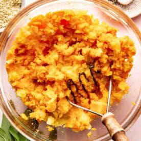 A glass bowl of mashed potatoes with a potato masher, surrounded by spices, salt, pepper, and fresh sage leaves on a countertop.