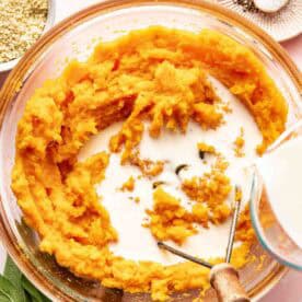 A bowl of mashed sweet potatoes with milk being poured in, surrounded by a bowl of breadcrumbs, fresh sage, and a plate of salt and pepper.