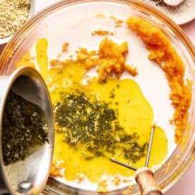 A glass bowl with mashed sweet potatoes, melted butter with herbs being poured in, and a masher beside it; salt, pepper, and breadcrumbs are nearby.