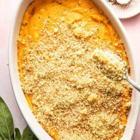 An oval white baking dish filled with sweet potato casserole topped with breadcrumb mixture; spoon adding more crumbs. A plate with salt and pepper and fresh sage leaves nearby.