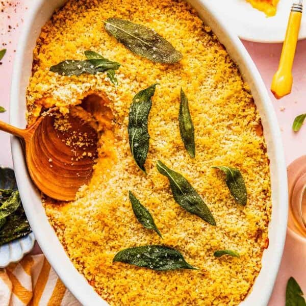 Baked savory sweet potato casserole with a golden breadcrumb topping and sage leaves, served in a white dish with a wooden spoon and a portion plated nearby.