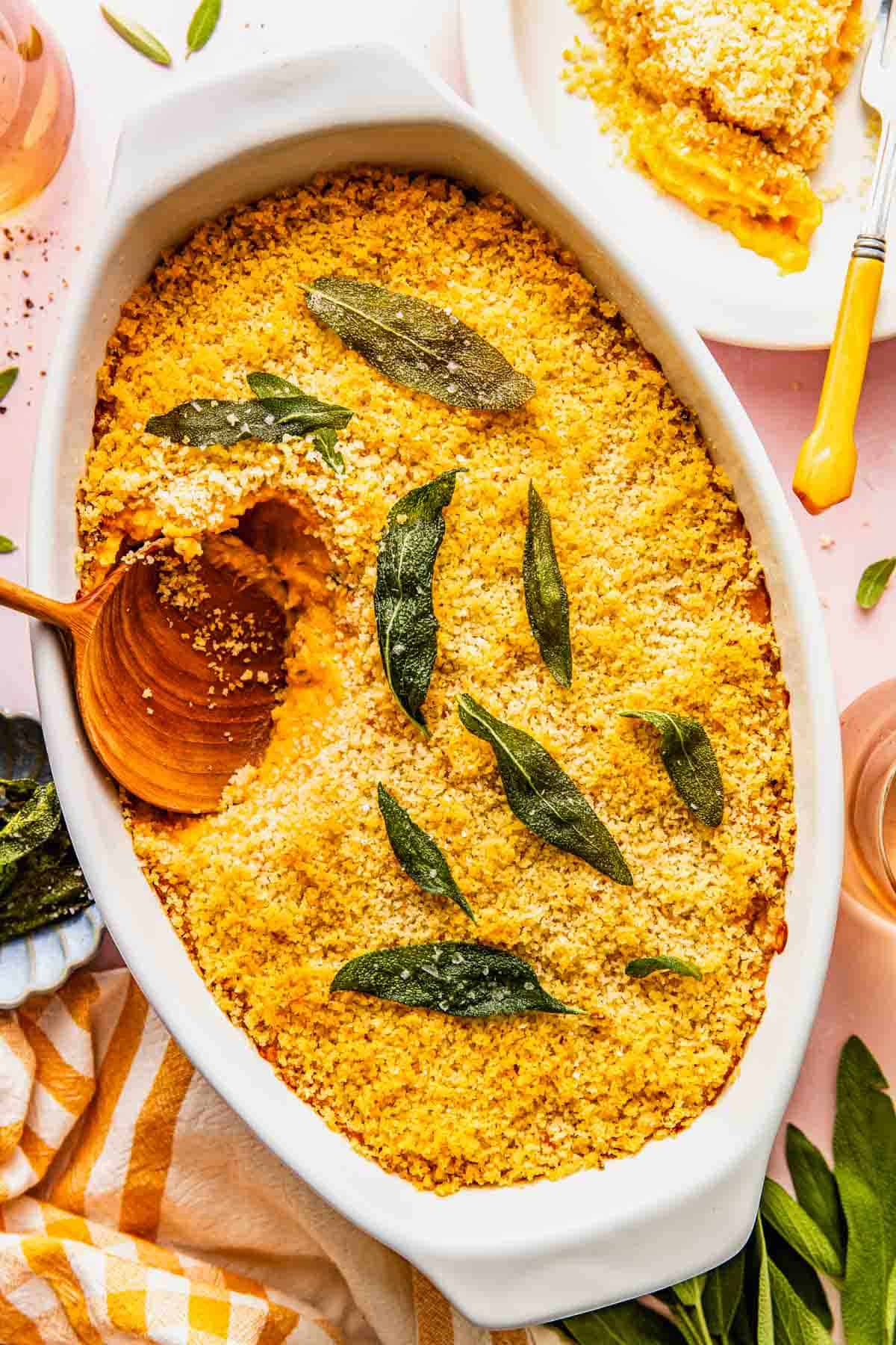 Baked savory sweet potato casserole with a golden breadcrumb topping and sage leaves, served in a white dish with a wooden spoon and a portion plated nearby.