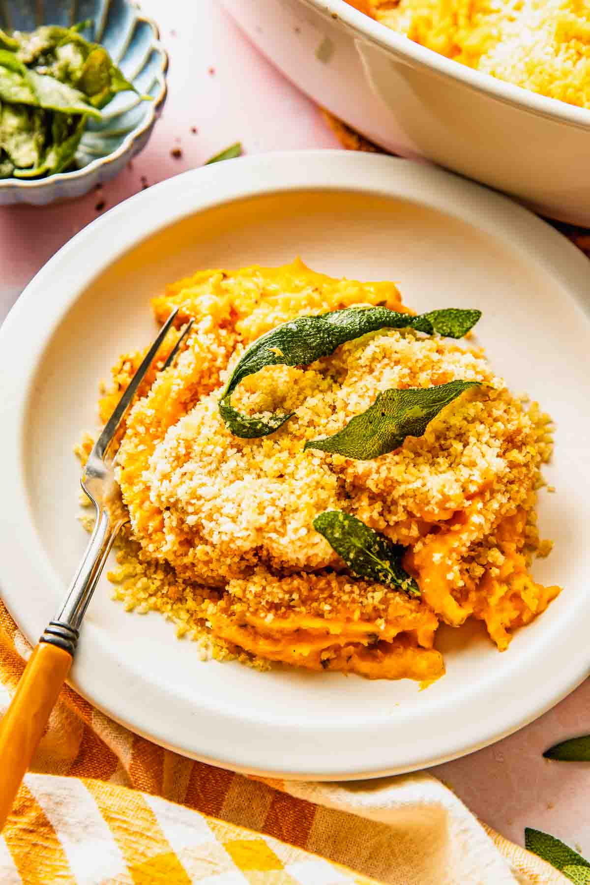 A plate of savory sweet potato casserole filling topped with breadcrumbs and crispy sage leaves, served with a fork on a white plate.