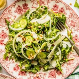 A salad with arugula, sliced cucumbers, onions, shaved cheese, and pine nuts is served on a decorative plate with a fork on the side.