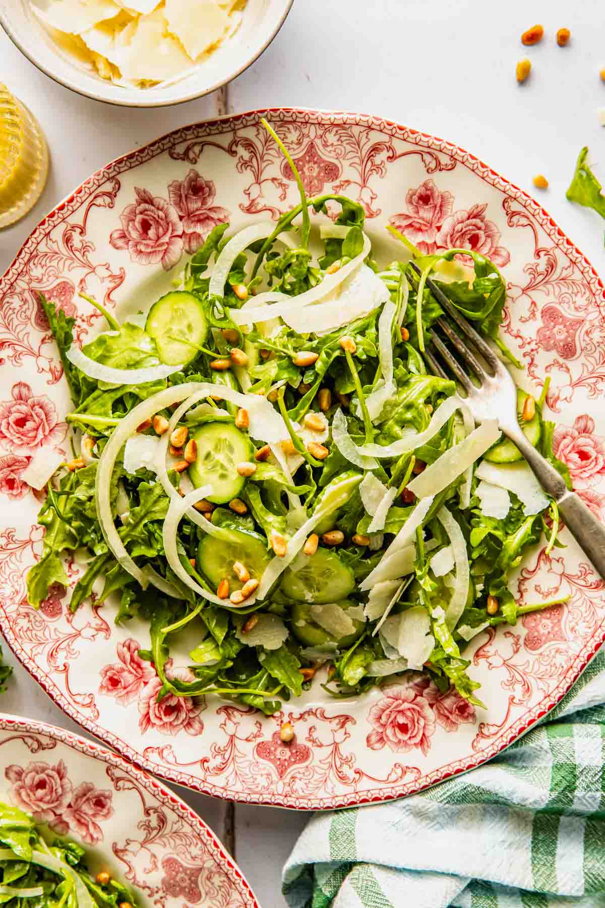 A salad with arugula, sliced cucumbers, onions, shaved cheese, and pine nuts is served on a decorative plate with a fork on the side.