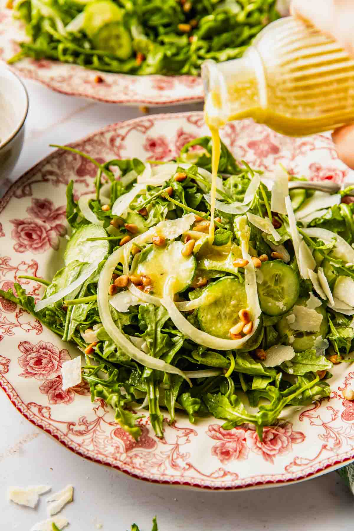 A hand pours dressing onto a salad with arugula, cucumber, shaved parmesan, fennel, and pine nuts on a floral-patterned plate.