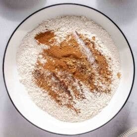 A bowl with flour and spices, surrounded by eggs in a bowl, a stick of butter, and a dish of dark liquid on a light surface.