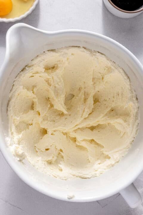 A white mixing bowl filled with creamed butter and sugar, with an egg and a small bowl of vanilla extract nearby on the countertop.