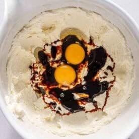 A mixing bowl with creamed butter and sugar, two raw eggs, and molasses on top, ready to be mixed.