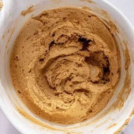 A white bowl containing light brown cookie dough being mixed, viewed from above on a light surface.