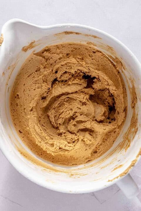 A white bowl containing light brown cookie dough being mixed, viewed from above on a light surface.