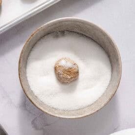 A cookie dough ball is coated in white granulated sugar in a small bowl, with more dough balls on a baking sheet and a scoop of brown sugar nearby.