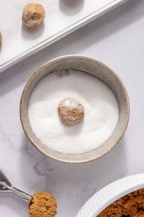 A cookie dough ball is coated in white granulated sugar in a small bowl, with more dough balls on a baking sheet and a scoop of brown sugar nearby.