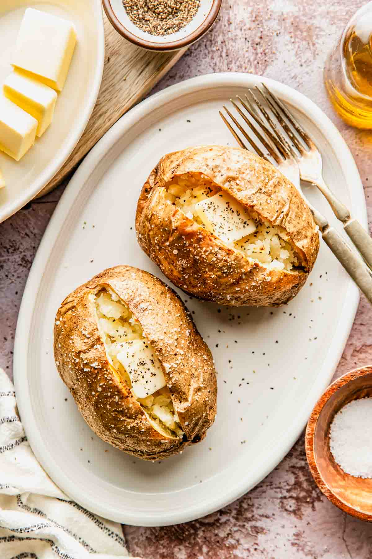Two baked potatoes with butter and black pepper on a white oval plate, accompanied by a fork and knife. Butter, pepper, salt, and a glass of oil are also visible.