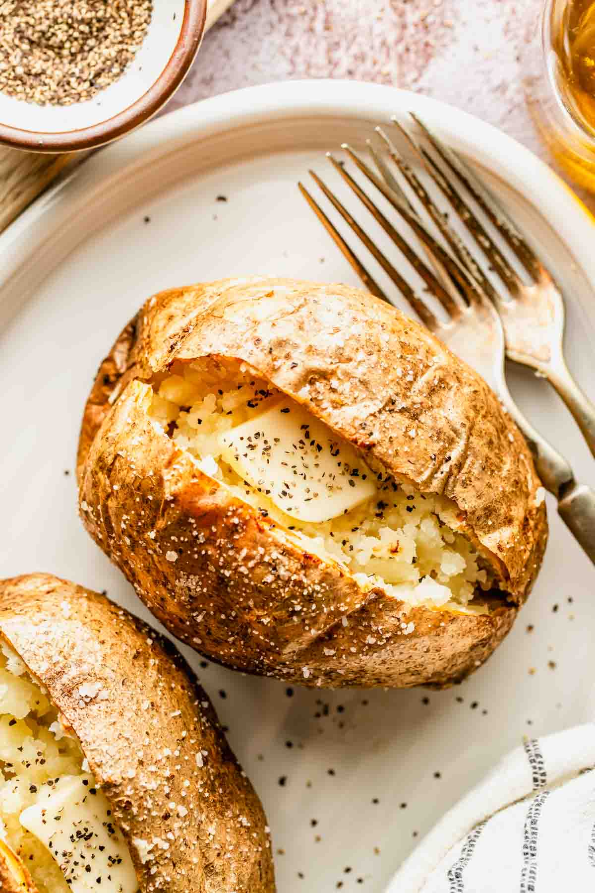 A baked potato split open on a plate, topped with butter and black pepper, with a fork and knife beside it.