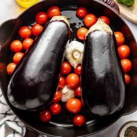 Two whole eggplants, cherry tomatoes, and a head of garlic in a cast iron skillet, surrounded by olive oil, fresh basil, salt, pepper, and a kitchen towel.