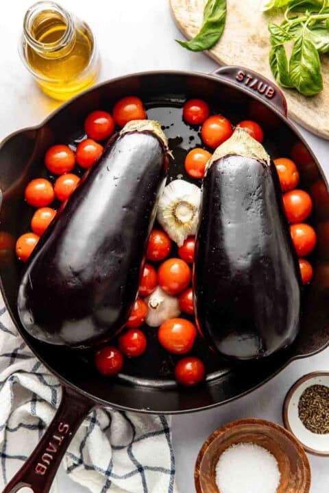 Two whole eggplants, cherry tomatoes, and a head of garlic in a cast iron skillet, surrounded by olive oil, fresh basil, salt, pepper, and a kitchen towel.